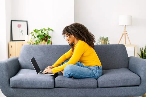 Smiling young woman with curly hair using laptop while relaxing on sofa at home Stock Photos