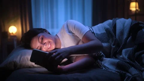 Smiling young woman lying in bed at night, scrolling through social media on her Stock Footage 305167862