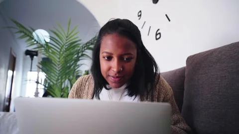 Smiling young woman using laptop sitting on sofa at home. African female in Stock Footage 252070880