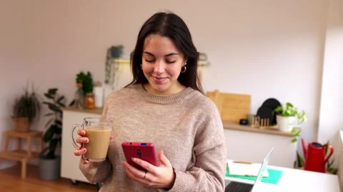 Smiling young woman using smart phone while drinking coffee at home Stock Footage 303429400