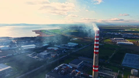 Smoke rises from a tall stack near factories by the water during daylight with a 库存影片 335359141