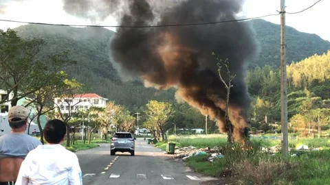 Smoke rising from a fire in a dumping site by the street while a car and people Video stock 143623115