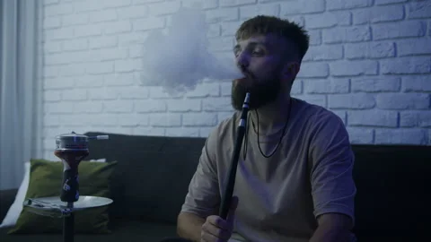 Smoking hookah at home. A man exhales smoke while sitting on the couch Stock Footage 204842353