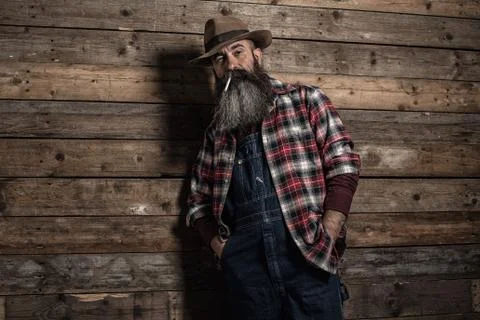 Smoking vintage worker man with long gray beard in jeans dungarees. Standing  Stock Photos