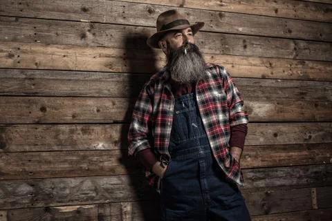 Smoking vintage worker man with long gray beard in jeans dungarees. Standing  Foto stock