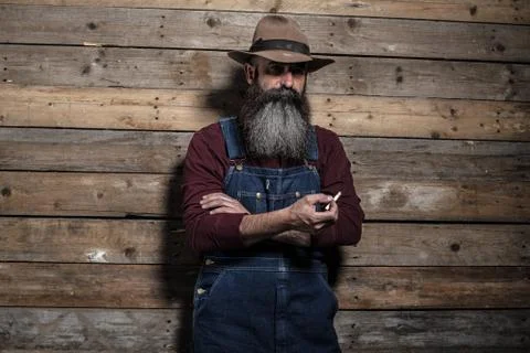 Smoking vintage worker man with long gray beard in jeans dungarees. Standing  Stock Photos