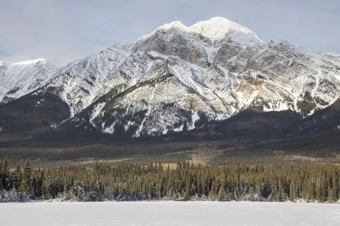 Snow-capped mountain range with evergreen forest and frozen lake during win.. Stock Photos