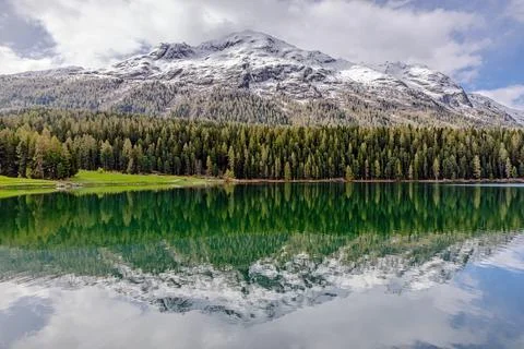 Snow-capped mountains reflect in tranquil lake surrounded by lush forest Stock Photos