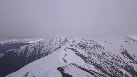 Snow covered mountain ridge with low clouds hiding peak. Ascending aerial Video stock 249054047