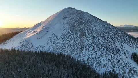 Snow-covered mountain rises above dense evergreen forest during sunrise Video stock 293083866