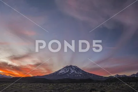 The snow-covered slopes of a volcano are set against a red sunset sky Stock Photos