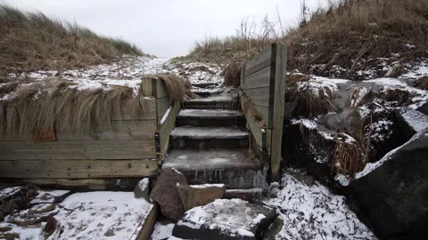Snow-covered steps lead up through grassy dunes under a cloudy winter sky Stock Footage 288474342