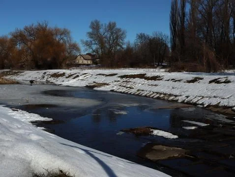 Snow covers the banks of a river while a house sits nearby under a clear blue 스톡 사진