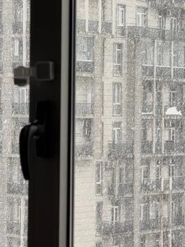 Snow falling outside a closed window with a view of an apartment building Stock Photos