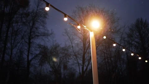 Snow falling on string lights hanging from a lamp post Stock Footage 291392616