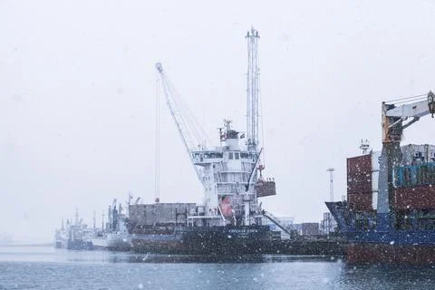 Snow falls during winter as cargo ships are docked at a port Stock Photos