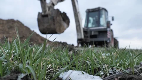Snow on green grass. On an blurry background, an excavatar digs a trench. Stock Footage 236775145