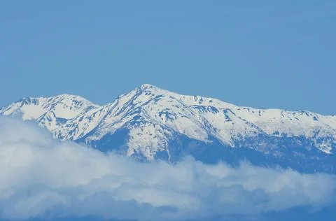 A snow-topped mountain range rises above white clouds into a blue sky. Stock Photos