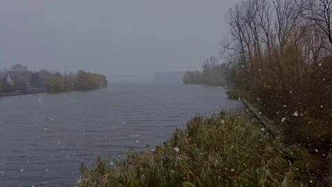 Snowing in a gray winter day by the lake. Large snow flakes over the lake. Video stock 258243422