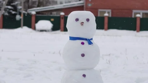 Snowman with blue scarf looking at camera against snowfall background in winter Stock Footage 314888285
