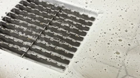 Soap bubbles and foam gather around grated floor drain in sanitary white sink Stock Footage 321942996