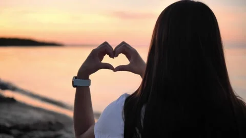 In the soft light of sunset on the seashore, young woman forms a heart with her Vidéo 282200994
