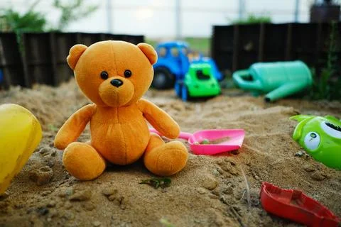 A soft orange teddy bear is placed in a sandbox with vibrant plastic toys s.. Stock Photos