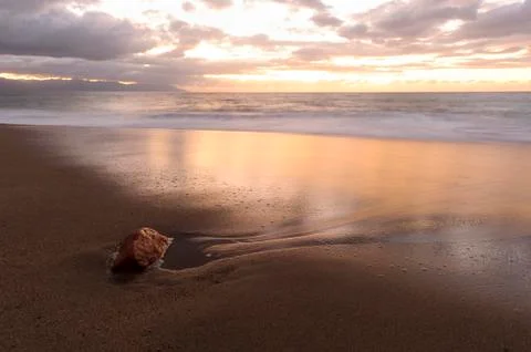 A soft sandy sunset with a cloud filled sky and a lone beach rock on the seashor Stock Photos