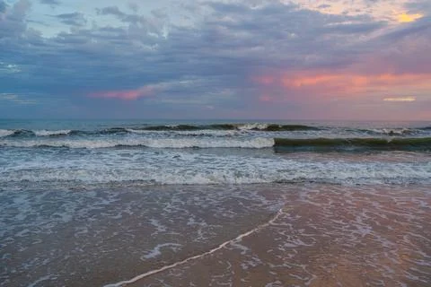 Soft waves roll onto the shore under a sky tinted with pink and purple hues. Stock Photos