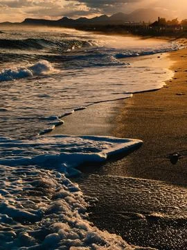 Soft waves washing sandy beach at sunset with mountains in background. Soft Stock-Fotos