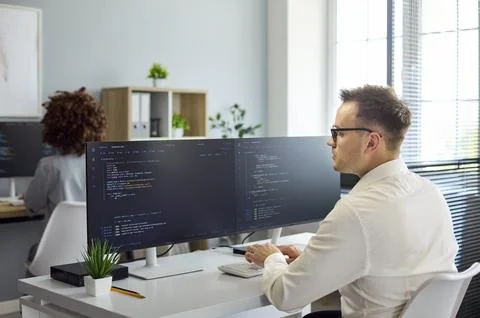 Software developer coding at dual monitors in modern office workspace, daylight 스톡 사진
