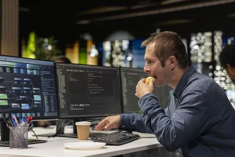 Software developer eating croissant in office, reviewing AI system metrics 库存照片