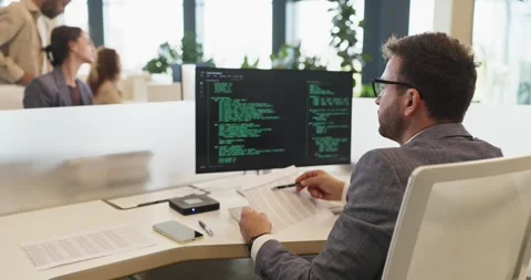 Software developer reviewing documents beside code monitor in modern office Stockbeeldmateriaal 347600354