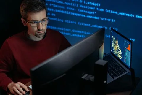 Software engineer in glasses sits in a dark room at a desk with multiple Stock Photos