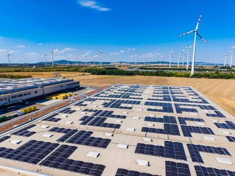Solar panels cover a large flat roof while wind turbines rotate in the back.. Stock Photos