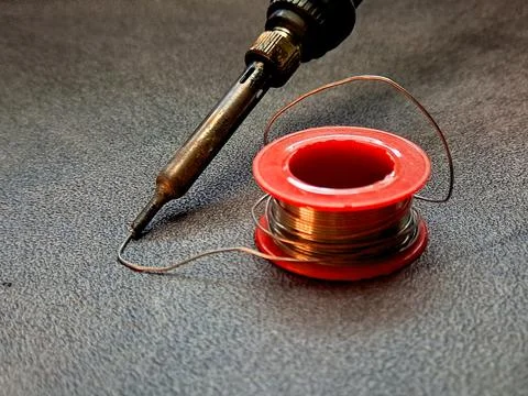 A soldering iron is placed next to a spool of copper solder on a textured s.. Stock Photos