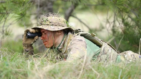 Soldier lies in wait, watching through field glasses, talking on radio. Stock Footage 127753589
