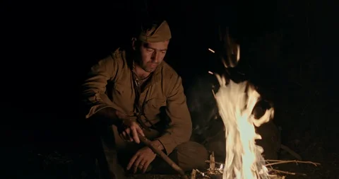Soldier in uniform crouching down on the ground stoking a blazing campfire at Stock Footage 128232514