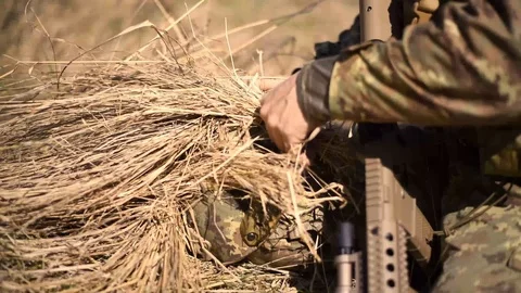 A soldier in a war scout puts dry grass in camouflage helmet to create Video stock 107388853