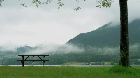 A solitary picnic table under a tree by a lake, with misty mountains in the Video stock 281592082
