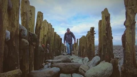 A solo hiker walks in between old rustic pier pillars on a beach Stock Footage 269690971