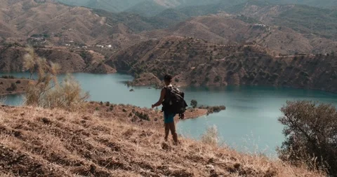 Solo man hiking down a dry grassy slope towards a vast blue reservoir in ar.. Stock Footage 340995945