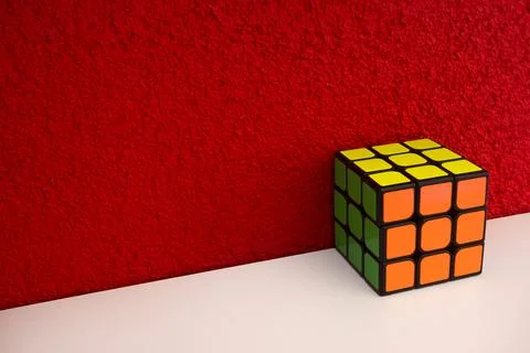 Solved puzzle speed cube on white wooden shelf near red colored wall, colorful Stock Photos