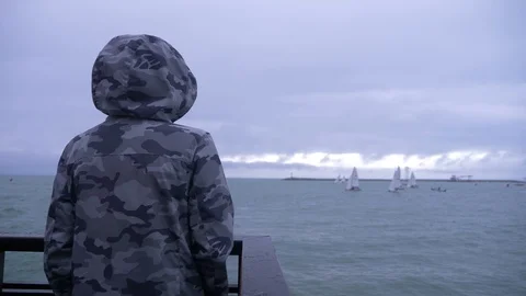 Someone in a hooded jacket is standing on the pier and watching the sailboats Видео 87905749