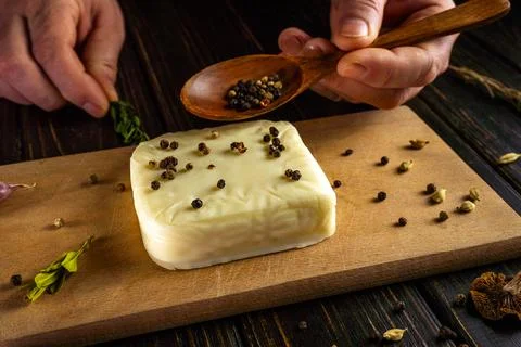 Someone is preparing cheese by adding spices to a block on a wooden surface.  Stock Photos