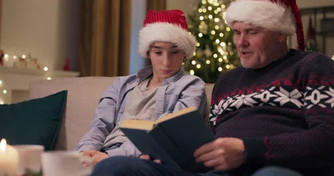 Son and dad sitting on couch dressed in Santa hats. Dad is holding book and Stockbeeldmateriaal 210623473