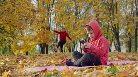 The son is playing on the phone while the mother is doing fitness in the park. Stock Footage 261835573
