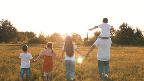 Son sits on father shoulders while family walks with across field at sunset Stock Photos