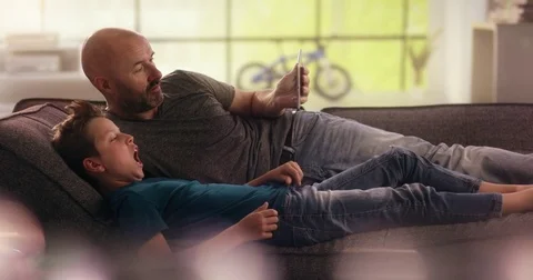 Son typing on the tablet his father is holding on the couch Video stock 78781055