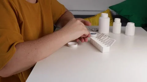 Sorting daily pills into white weekly pill organizer on home table with med.. Stock Footage 318456665